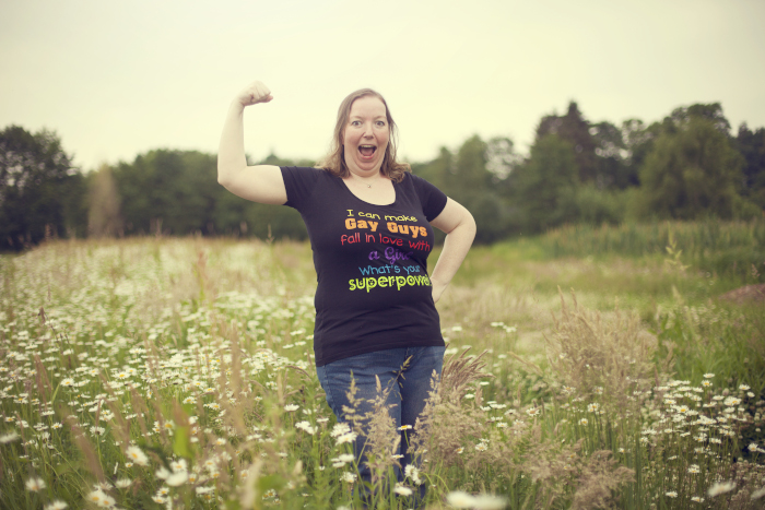 Pauline in een bloemenveld met haar arm omhoog om haar spierballen te laten zien. Tekst op het shirt in tegenboogkleuren: "I can make gay guys fall in love with a girl, what's your superpower?"